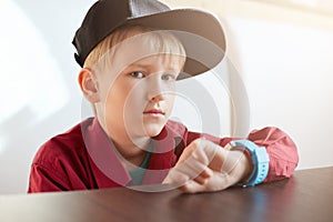 A horoizontal portrait of serious male child wearing trendy cap and red shirt having a smart watch on his wrist sitting at wooden