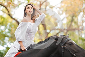 Horseback riding. Beautiful young woman in a white dress riding on a brown horse outdoors.