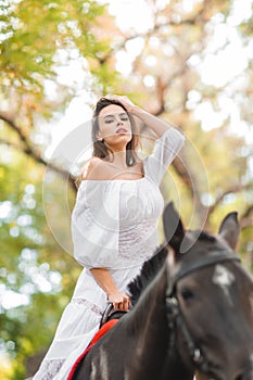 Horseback riding. Beautiful young woman in a white dress riding on a brown horse outdoors.