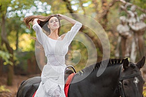 Horseback riding. Beautiful young woman in a white dress riding on a brown horse outdoors.