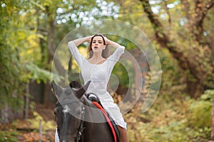 Horseback riding. Beautiful young woman in a white dress riding on a brown horse outdoors.