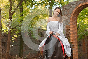 Horseback riding. Beautiful young woman in a white dress riding on a brown horse outdoors.