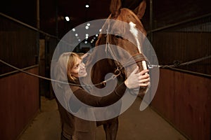 Horsewoman petting horse stroking animal muzzle over stable interior