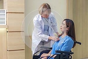 In a hospital hallway, a smiling doctor examines a patient, an Asian woman paralyzed and seated in a wheelchair