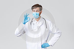 Hospital, healthcare workers, covid-19 treatment concept. Cheerful doctor in white scrubs, gloves, medical mask, okay