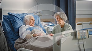Hospital Ward: Elderly Man Resting in Bed, His Caring Beautiful Wife Supports Him Sitting Beside,
