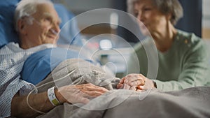 Hospital Ward: Focus on Hands of Elderly Man Resting in Bed, His Caring Wife Supports Him By Sitti