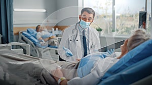 Hospital Ward: Friendly Doctor Wearing Face Mask Talks to Beautiful Senior Female Patient Resting in