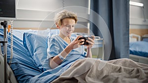 Hospital Ward: Handsome Young Boy Resting in Bed, Uses Smartphone, Plays Online Video Games on Int