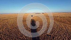 Hot air ballon traveling over dry grass field with shadow on ground