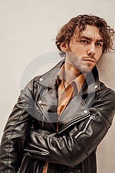 Daring and cool male model with curly hair posing in leather coat in a bright studio