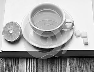 Hot beverage with sugar cubes placed on open book on wooden background. Cup of green tea with slice of lemon.