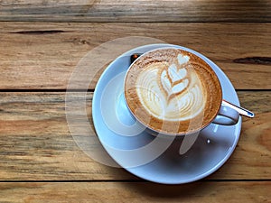 Hot cappuccino coffee in white cup and saucer with spoon on wooden table background. Art of milk foam drawing.