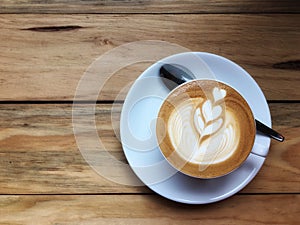Hot cappuccino coffee in white cup and saucer with spoon on wooden table background. Art of milk foam drawing like white heart on