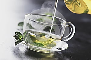 Hot chinese green tea with mint, with splash pouring from the kettle into the cup, steam rises, dark background, selective focus