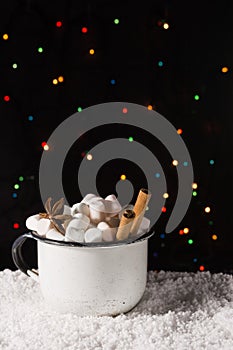 Hot chocolate in cup with marshmallow and spices on the snow with christmas lights background. Copy space