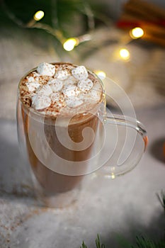 hot chocolate in a glass mug with mini marshmallows on grey table, blurred lights background