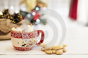 Hot chocolate with marshmallow candies. Christmas cookies shaped in snowflakes, golden cones and christmass tree lights. White wo