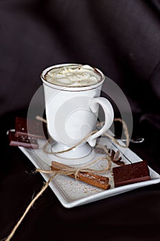 Hot chocolate in a mug with handle, spices and chocolate pieces on white plate black background