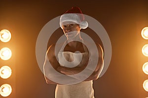 Hot Christmas dessert. Muscular man wearing apron and Santa hat folded arms and smiling at camera, lamps illumination on