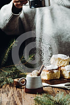 A hot cocoa with cinnamon stick and fresh Christmas buns with powder and over festive table, a girl is sprinkling sugar powder on