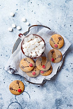 Hot cocoa with marshmallow in ceramic mug surrounded by Christmas gingerbread. Decorated red nosed reindeer cookies.