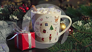 Hot cocoa with marshmallows, cinnamon in white mug, surrounded by winter Christmas tree branches with berries, red gift box and