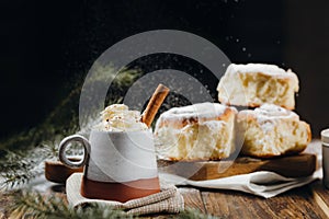 A hot cocoa with whipped cream, cinnamon stick and fresh Christmas buns with powder and over festive table, sugar powder is