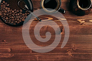 Hot coffee in black cup with beans, spoon and turkish pot cezve with copy space on brown old wooden board background, top view.