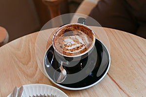 Hot coffee with latte art in black ceramic cup on wooden table. Cappuccino or latte with frothy foam milk.