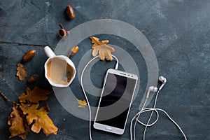 Hot coffee in mug and bun, mobile phone with headphones and autumn leaf