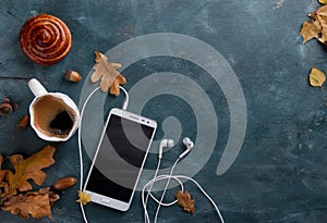 Hot coffee in mug and bun, mobile phone with headphones and autumn leaf