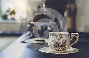 Hot cup of tea with blurry cake on hight tea stand in the kitchen background. Cup of aroma dring with steam, Cozy sence of