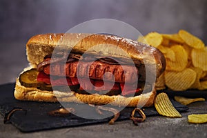 Hot dog -Hot sausage nested in a bun with cucumbers, red pepper and onions. On a dark background with chips. Fast food concept