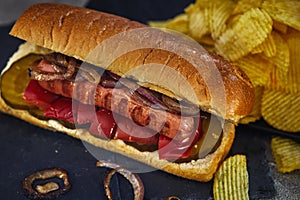 Hot dog -Hot sausage nested in a bun with cucumbers, red pepper and onions. On a dark background with chips. Fast food concept