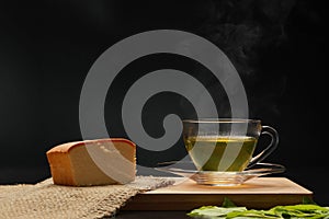 The hot green tea in the glass cup with smoke and butter cake on the wooden board as black background. Healthy drink concept. Dark