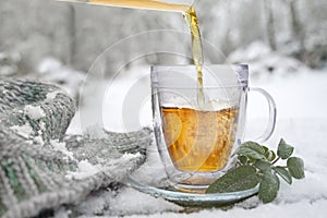 Hot herbal tea is poured in a glass cup outdoors in the snow, beside some sage leaves and a knitted scarf, medicinal herbs and