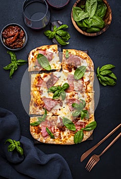 Hot homemade roman pizza with ham, mozzarella cheese, sun dried tomatoes, sauce and green basil, black table background, top view