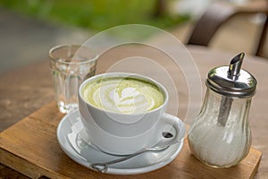 Hot Matcha Latte in white cup with drink water in glass and sugar in bottle on wood table