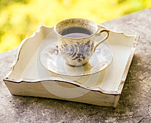 Hot morning coffee in small blue and white floral design cup on shabby chic vintage white tray on window sill in front of summer