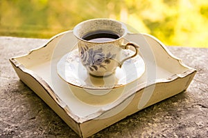 Hot morning coffee in small blue and white floral design cup on shabby chic vintage white tray on window sill in front of summer