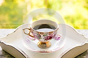 Hot morning coffee in small floral design cup with gold on shabby chic vintage white tray on window sill in front of summer window