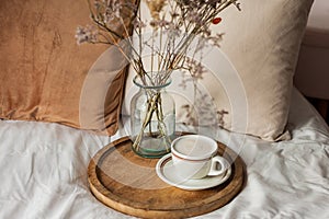 Hot mug of cappuccino on wooden tray on the bed, breakfast, bouquet of dried flowers. Spring scenery. Cozy house. Beige natural