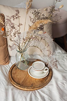 Hot mug of cappuccino on wooden tray on the bed, breakfast, bouquet of dried flowers. Spring scenery. Cozy house. Beige natural