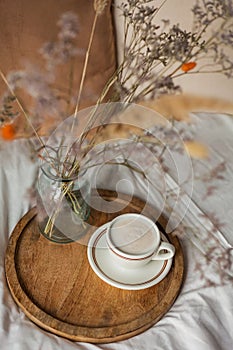 Hot mug of cappuccino on wooden tray on the bed, breakfast, bouquet of dried flowers. Spring scenery. Cozy house. Beige natural