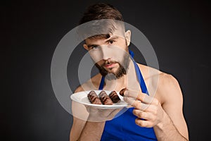 Hot naked handsome man with chocolates, chocolate candies. Smiling naked handsome man wearing dark blue apron holding a white