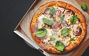 Hot pizza in box with spicy salami sausage, mozzarella cheese, tomato sauce and green basil, just delivered, black table