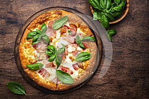 Hot pizza with ham, pancetta, mozzarella cheese, tomato sauce and green basil, rustic wooden table background, top view