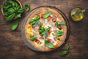 Hot pizza with ham, pancetta, mozzarella cheese, tomato sauce and green basil, rustic wooden table background, top view