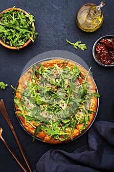 Hot pizza with mozzarella cheese, ham, pesto and fresh arugula, black table background, top view